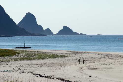 To mennesker vandrer på en lang, vid strand med spisse fjellformasjoner i bakgrunnen.