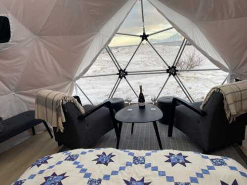Inne i arctic dome med dobbeltseng, to lenestoler og et lite bord med vinflaske og glass.