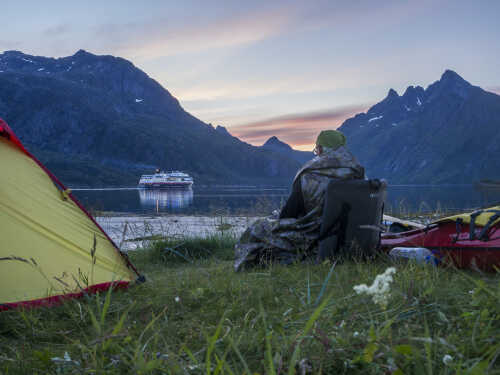 Mann sitter i gresset ved teltet med kajakken ved siden av seg og ser utover fjorden på hurtigruten.